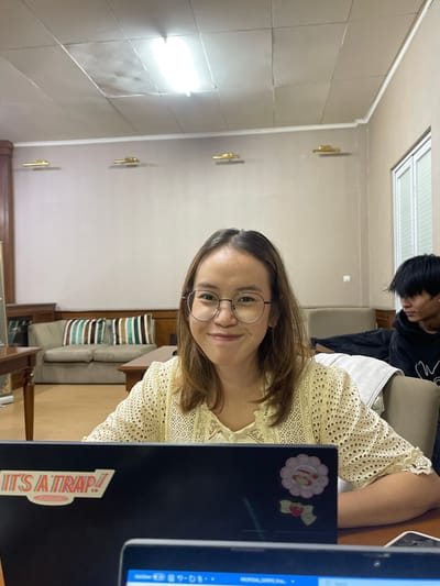 The image depicts an indoor scene in Muka, Indonesia, featuring two individuals in what appears to be a common room or study area. A young East Asian female, wearing round spectacles and a cream-colored, short-sleeved eyelet top, occupies the foreground, smiling directly at the viewer. She is seated at a wooden table behind a black laptop displaying "IT'S A TRAP!" and decorative stickers. A white smartphone with a cracked screen is on the table to her right. In the background, positioned to the right of the frame, a young East Asian male with dark hair and a dark hoodie is seated, facing away from the camera. The room's environment includes light beige walls, a paneled ceiling with a central fluorescent light fixture, and three golden-toned wall sconces. A beige two-seater sofa with three striped cushions (green, dark brown, light brown) and a dark wooden table are visible on the left. A window with white louvered blinds is present on the right wall.