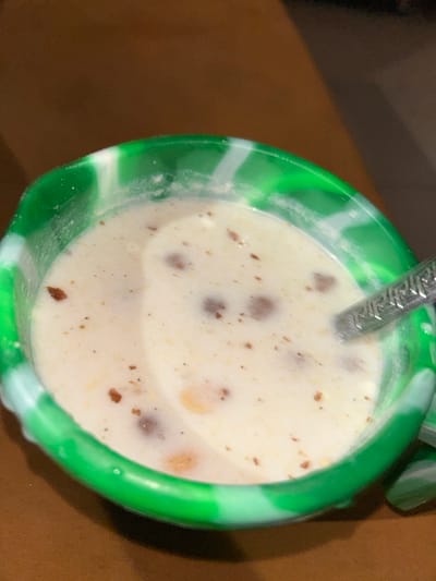 A close-up, top-down view depicts a bowl containing a liquid food substance within an indoor setting in Egbe, Nigeria. The bowl is circular, made of a green and white marbled material, possibly silicone. It holds a light, creamy-colored liquid in which various solid components are suspended: numerous dark brown, irregularly shaped spherical particles, smaller lighter brown/orange fragments, and fine dark specks. A silver-colored spoon, featuring a decorative pattern on its handle, is partially submerged in the liquid on the right side of the bowl. The bowl rests on an out-of-focus, dark brown surface, likely a table. No individuals or actions are visible.