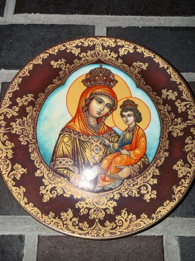 A decorative plate, depicting a religious icon of the Virgin Mary and the Christ Child, is displayed on a dark, textured brick surface. The plate features intricate gold scrollwork on a deep red background. The central image shows Mary wearing a crown and orange veil, holding the Christ Child who is also crowned and dressed in orange. Both figures have halos. The plate rests on a wall composed of alternating dark grey and lighter grey bricks, with visible mortar lines. This location is in Heiligenrode, Germany.