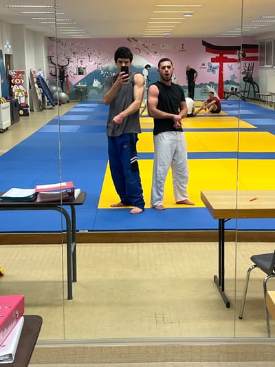 Two men stand on a blue and yellow judo mat, reflected in a mirror. One wears a black shirt and white pants, the other a gray shirt and blue pants. The second man holds a phone, seemingly taking a selfie. In the background, a wall features a mural with Japanese-inspired art, while workout equipment is visible along the wall. A table and some office supplies are also in the shot.