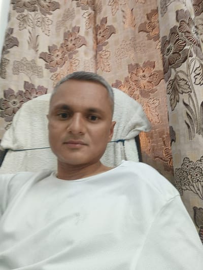 A man with short, graying hair and fair skin is pictured in a close-up, head-and-shoulders shot. He is wearing a white t-shirt and is seated in a chair. The background features a patterned curtain with floral motifs in shades of brown and beige. The man is looking directly at the camera. The location is Dhandhuka, India.