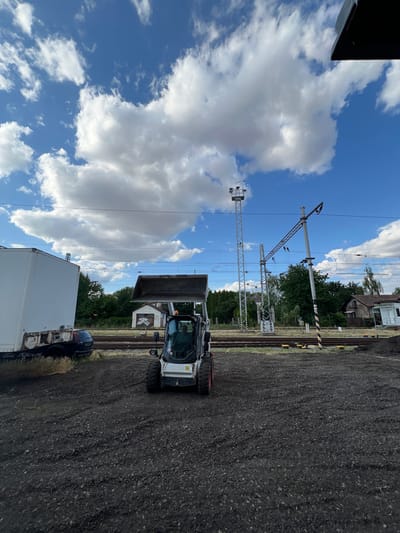 The image depicts a scene near a train track in Újezd u Brna, Czechia. A white skid steer loader with a raised bucket is positioned on dark, loose ground in the foreground. A white box truck is parked to the left of the skid steer. In the background, train tracks run horizontally, flanked by trees and a few small buildings. Power lines and support poles are visible, indicating an electrified railway line. The sky is blue with scattered white clouds. There is a person visible in the driver's seat of the skid steer.