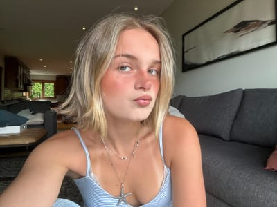 A young blonde woman is depicted in a close-up, waist-up, indoor portrait, likely a selfie, within a residential setting in Squamish, Canada. She has light-colored, shoulder-length hair and fair skin with visible freckles on her nose and cheeks, accentuated by noticeable pink blush. Her eyes are grey-green, and she has a silver hoop nose ring. Her lips are slightly parted in a pout, covered with a clear or light pink gloss. She is wearing a light blue and white striped camisole with white lace trim on the neckline, and a distinct tan line is visible across her chest and shoulders. She wears layered silver necklaces; one is a finer chain with small green and white beads and a small charm, and the other is a longer chain with a prominent, textured silver starfish pendant.

The background reveals a spacious open-concept room, dimly lit but with natural light entering from windows. To the left, a kitchen area is visible with dark wooden cabinetry, a dark countertop, a sink with a faucet, and a large window looking out onto lush green trees. Recessed lighting is visible on the ceiling. To the right, a living room area features a dark grey fabric sofa with textured upholstery. Above the sofa, a large, horizontal framed abstract artwork is mounted on a light-colored wall, featuring hues of white, black, and earthy brown/orange. In the mid-ground, a section of a brown leather or upholstered bench/ottoman is visible, along with parts of blue and white pillows and a dark rug on the floor.