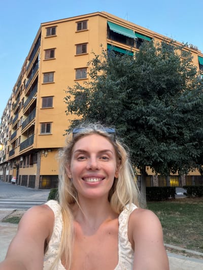 A young woman with blonde hair takes a selfie in front of a yellow apartment building. She is smiling and looking at the camera. The building has multiple stories with many windows, some with wooden shutters and others with green awnings. A large tree with dark green foliage is positioned in front of the building, partially obscuring it. The sky is clear and blue, suggesting it is daytime. The woman is wearing a white, lacy top. The overall scene appears to be an outdoor urban or suburban environment. The image is oriented vertically, with the woman's head at the top and the building extending downwards. There is no visible text in the image.