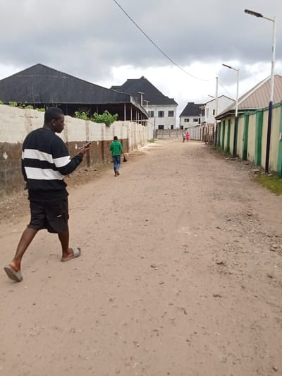 The image shows a dirt road in Ovwian, Nigeria, located at 5.485 latitude and 5.806 longitude. Three people are walking down the road, two are in the distance and one is closer to the camera, walking away from the viewer. The person closest to the camera is looking at a mobile phone.  The road is lined with houses and buildings on both sides, and there are several power lines and light poles running along the road. The houses are two-story and appear to be relatively new, with some having a white facade and a corrugated metal roof. The ground is dirt and covered in small stones. The sky is overcast with clouds.