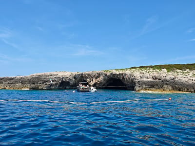 The image depicts a scenic view of the sea near the city of Grad Vis, Croatia. A rocky cliff face with caves dominates the background, with green vegetation growing on top. In the foreground, the deep blue sea is rippled by gentle waves. A rope with buoys stretches across the water, marking a boundary. Two boats are anchored near the cliffs, one larger with several people visible on deck, and a smaller dinghy. The sky is clear and blue with a few wispy clouds. The overall impression is one of a bright, sunny day in a beautiful coastal location.