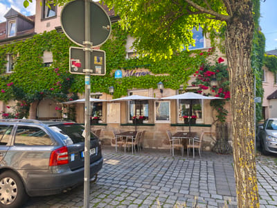 This media file depicts an outdoor seating area of a restaurant or cafe in Erlangen, Germany. A gray station wagon is parked on a cobblestone street in the foreground, partially obscuring the view of the establishment. The building is covered in lush green ivy, with vibrant pink roses climbing its facade. Several tables and chairs are arranged outside, covered by white umbrellas, suggesting an inviting atmosphere for dining or relaxation. Large windows offer a glimpse into the interior. A tall tree with textured bark stands prominently on the right side of the image, its leaves dappled with sunlight, indicating it is daytime. The sky is visible and appears to be a clear blue with some clouds. A street sign with various stickers and a circular metal sign are mounted on a pole in the left foreground. The overall impression is a pleasant, possibly sunny day, with the restaurant's greenery and flowers creating a charming and picturesque scene.
Visible text includes "Marino" on the building facade, a license plate on the car, and various stickers on the street sign. The license plate appears to be "F 959", and a blue sticker with a white wheelchair symbol is also visible. The stickers on the street sign are too small to be legible.