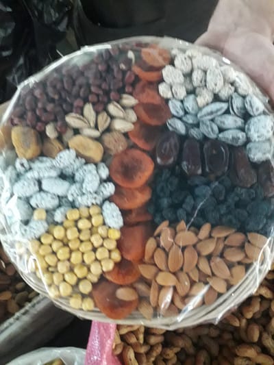 A hand holds a circular platter, wrapped in clear plastic, showcasing an assortment of dried fruits and nuts. The platter is segmented into distinct sections, each filled with a different item.

Starting from the top left and moving clockwise, the segments include: light-colored, possibly sugar-coated dried fruit or nuts; dark, shriveled fruits such as dates, followed by dark raisins; light brown, oval-shaped almonds; bright orange dried apricot halves; and round, yellow roasted chickpeas (likely "nohot"). The bottom left section contains small, dark red items, possibly dried berries or small nuts. A central segment at the bottom also contains light-colored, sugar-coated items, resembling frosted nuts or chickpeas. Interspersed among these are more dried apricots and some other light-colored nuts or seeds.

The overall scene suggests a market setting, likely in Ghafurov, Tajikistan, given the common presentation of such assortments in Central Asian markets. In the background, large bulk bags or containers are visible, filled with unshelled peanuts and other unidentified nuts or dried goods, reinforcing the market stall environment. A piece of pink plastic or fabric is also partially visible in the upper right background. The image is slightly blurry, but the variety of items on the platter is clear. No text or specific time of day is discernible.