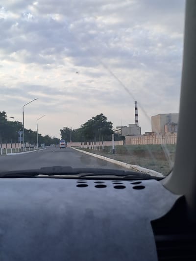 The image is a point-of-view shot from inside a vehicle, looking out at a road leading towards industrial buildings. The sky is overcast with light clouds. On the right side of the road, there is a large, light-colored industrial building with a tall, red and white striped smokestack. A fence runs along the front of the industrial complex. On the left side of the road, there are streetlights, trees, and a sidewalk. A white bus is visible driving away on the road, and other vehicles and people can be seen further down. The foreground of the image shows the dashboard of the vehicle. The scene appears to be located in an industrial area on the outskirts of a town or city.