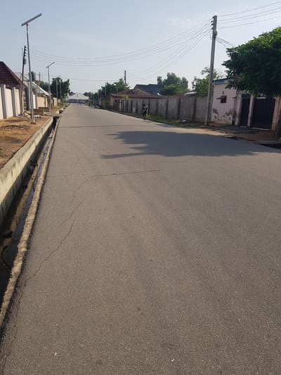 The image depicts a street scene in Palade, Nigeria. The perspective is from the side of the road, looking down its length. The road itself is paved with asphalt and shows some signs of wear, with visible cracks. Along the left side of the road, there is a raised concrete curb and a drainage ditch, which appears to contain some water and debris. Beyond the curb, a dusty embankment is visible, with some scattered litter.

On the left side of the street, a utility pole with numerous wires stretches upwards, disappearing out of frame. In the background, various buildings and structures line the road. Some appear to be residential houses, with plastered walls, some of which are weathered and discolored. There are also trees with lush green foliage scattered throughout the scene. Further down the road, more buildings and more utility poles with overhead wires are visible. There are also solar-powered streetlights positioned along the road.

A person on a motorcycle can be seen in the distance, traveling down the street. The sun is high in the sky, casting long shadows across the road, suggesting it is daytime, likely late morning or early afternoon. The sky is clear and bright, indicating fair weather. The overall impression is of a typical urban or suburban street in a tropical climate. There is no visible text in the image.
