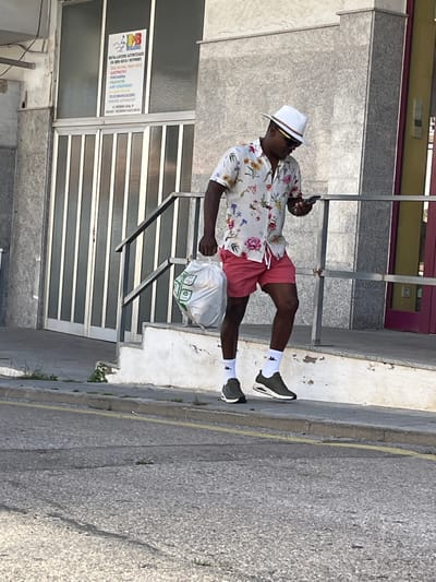 A man is walking on a sidewalk in Calafell, Spain. He is wearing a white hat, sunglasses, a white floral shirt, red shorts, white socks, and black shoes. He is carrying a white plastic bag. The sidewalk is gray and the street is asphalt. There is a building in the background with a sign that says "RESTA. LACIONE AUTORIZADAS." The building has white shutters.