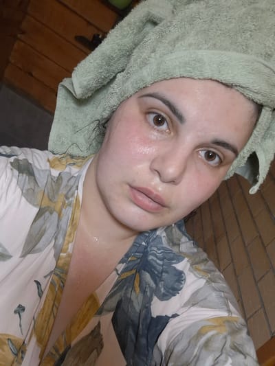 A close-up, tilted selfie of a person with a fair complexion and light brown eyes. The person has a green towel wrapped around their head. Their skin appears slightly flushed and possibly damp. They are wearing a robe or top with a floral print, including shades of green, blue, and yellow. The background shows a wooden floor or wall paneling. The location is unknown and cannot be determined from the image. Амзя, Russia cannot be identified as the city within this image.
