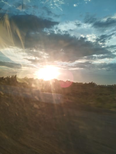 This image captures a sunset viewed through a window, likely from a moving vehicle, as indicated by the streaking effect in the foreground and the reflection on the glass. The sun is bright and low on the horizon, casting a warm glow and creating lens flares. Dark clouds are scattered across a blue sky, with some sunlight breaking through. The landscape in the background appears to be rural, with a line of trees or foliage along the horizon and a grassy field below. A road and a barrier are visible in the foreground, blurred by motion. The location is identified as Elabad, Uzbekistan.