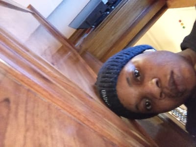 The image, oriented approximately 90 degrees counter-clockwise, depicts an indoor scene. A single individual, an adult with dark skin, occupies the central frame, wearing a dark knit beanie and a dark top, looking directly at the camera with a neutral expression.

To the left (when upright), warm-toned wooden paneling or a staircase banister is visible, comprising multiple parallel elements. To the right, a light-colored wall serves as the background. Mounted high on this wall is a dark, rectangular object. Below and to the upper right of the individual's head, a small item displaying three distinct colored circular indicators (red, orange, green) is affixed. A partially visible object with reflective surfaces is also present in the lower right background.

The scene is reportedly located in an interior setting in Madinaty, Egypt, though no specific visual identifiers within the image itself confirm this geographic location.