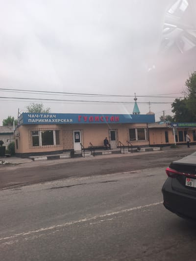 The image depicts a one-story building with a blue awning that reads "ЧАЧ-ТАРАЧ ПАРИКМАХЕРСКАЯ" and "ГУЛИСТАН". The building is peach colored with black trim. A man is sitting on a bench in front of the building. In the background, there is a church steeple. In the foreground, there is a road with a black car parked on the right side of the frame. The car has a KG license plate with the number 08. The location is Ленин, Kyrgyzstan. The sky is overcast.