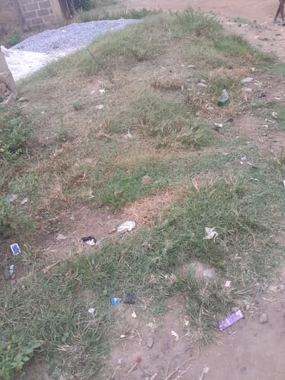 The image depicts an outdoor scene in Ifo, Nigeria, characterized by a mix of natural ground cover and discarded items. The foreground and midground consist of uneven terrain featuring patches of green and dry brown grass interspersed with exposed dirt and small stones. Scattered extensively across these areas are various pieces of litter, including multiple plastic wrappers (identifiable colors: purple, blue, white, black), what appears to be a green plastic bottle, and other unidentifiable debris.
On the left side, towards the mid-ground, a large pile of light grey aggregate (gravel) is visible on a dirt patch. Adjacent to this pile, to the left, is a quantity of white material, possibly sand or cement. Behind these materials, partial views of a building constructed from grey concrete blocks are discernible.
In the upper right portion of the frame, the lower legs and feet of a person are visible, walking away from the camera on a dirt path. The person is wearing light blue open-toed footwear. The overall environment appears to be an unpaved area, potentially a roadside or undeveloped lot within a residential or transitional zone, with signs of human activity and waste accumulation. The lighting suggests daylight conditions.