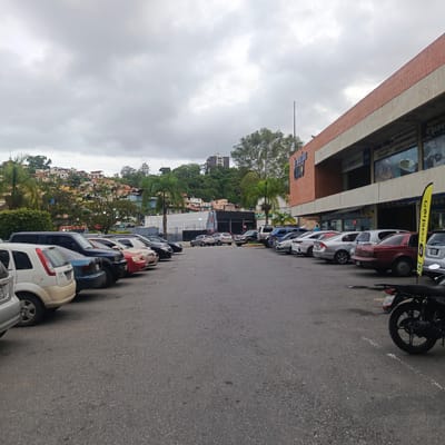 This is a medium shot, eye-level outdoor photograph taken on an overcast day, depicting a parking lot in front of a commercial building in Los Teques, Venezuela. The foreground is dominated by an asphalt parking lot with visible white lane markings. Several cars are parked in a line, including a white compact car on the far left, a dark blue SUV, a red sedan, and other vehicles further down the row. A black motorcycle is parked in the lower right corner of the frame.
In the mid-ground, palm trees and lush green vegetation are visible. Behind the vegetation, a hillside is densely populated with colorful houses, a characteristic of the urban landscape of Los Teques. The commercial building in the background has a brick facade on the upper portion and concrete trim, with large windows showcasing displays and signage for businesses. One sign reads "PRODUCTOS DISV" and another has the word "INTER" repeated.
The sky is heavily overcast with gray clouds, suggesting a lack of direct sunlight and potentially a humid climate. The overall impression is of a typical commercial area in a Venezuelan city, with a mix of residential and business elements. No people are clearly discernible in the image.