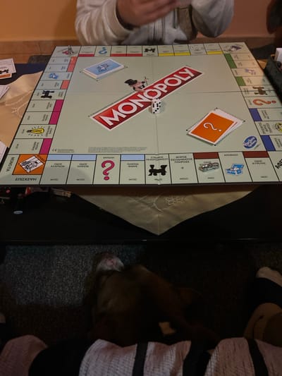 The image displays an indoor scene in the city of Gerakas Municipal Unit, Greece, centered on a Greek-language Monopoly board game. The board, featuring a prominent red and white "MONOPOLY" logo, is laid out on a dark table adorned with a light-colored, embroidered tablecloth. Two white dice, showing faces with one and two pips respectively, are positioned near the board's center. A silver-colored car token rests on a light blue property space, and another silver-colored token is located on a corner space. Stacks of blue (Chance) and orange (Community Chest) game cards are visible on their designated areas. In the upper portion of the frame, the hands and forearms of an individual, dressed in a light grey long-sleeved garment, are visible, engaged in holding and arranging game cards. The background features a plain, light-toned wall. In the lower foreground, beneath the table, a dark-furred dog, partially obscured, is lying on a dark carpet, with its snout visible. Adjacent to the dog, the legs of another individual, wearing striped socks or slippers, are present.