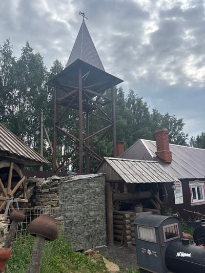 The image depicts an outdoor scene at what appears to be a rustic park or museum in Bol'shoi Mashlyak, Russia. In the foreground, on the left, is a large wooden water wheel. To the right of the water wheel, a tall, dark metal tower structure with a peaked roof topped with a weather vane rises, surrounded by trees. Attached to the base of the tower is a small building with a tin roof and brick chimney. In front of this building is a log structure with a well mechanism. Further to the right, a long, dark building with a corrugated tin roof and a prominent brick chimney is visible. A sign with Russian text is affixed to this building near a window with decorative trim. In the lower right of the image, a small, dark, train-like structure with windows and text that appears to be in Russian is present. The ground is a mix of gravel and grass, and there are stacks of stone and pottery-like objects in the scene. The sky is overcast with gray clouds. No people are visible in the image.