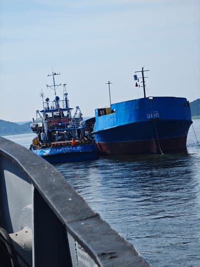 Two ships are docked together on a body of water. The ship on the left is a blue and white tugboat with "ПЛОТОBOД 717" printed on its side. A person wearing an orange life vest is visible on the deck of the tugboat. The ship on the right is a larger blue vessel with a red waterline. Both ships appear to be in the city of Podporozh'e, Russia, as indicated by text on the tugboat which reads "КРАСНОЯРСК" (Krasnoyarsk) and appears to be a port of registration. The water is calm with a slight ripple. The sky is clear and blue. In the foreground, a portion of another gray structure, possibly part of a bridge or another vessel, is visible. The background shows a tree-covered hill.