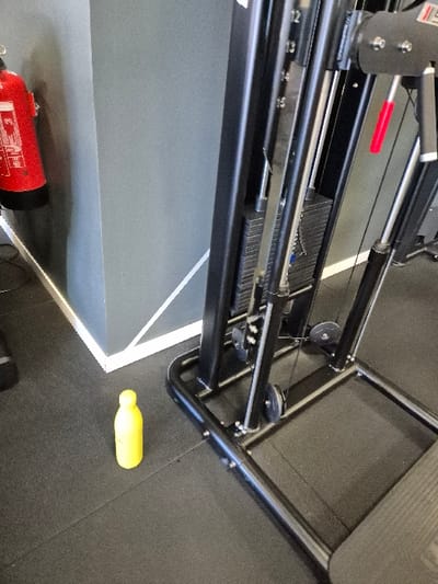 This image depicts an indoor fitness facility, presumably a gym, located in San Lorenzo de El Escorial, Spain. The scene is static, containing no visible people or discernible actions.

The foreground presents a dark, textured rubber floor. A bright yellow, opaque plastic bottle stands upright on this floor, slightly to the left of the image center.

To the right, a prominent black metal gym machine is positioned, featuring a visible weight stack with numerical indicators (e.g., 12, 13, 14, 15), cables, and multiple pulleys. The machine's base is constructed from black tubular metal, and a red handle is visible on a moving component towards the upper right section of the equipment.

To the left, two grey-painted walls form an interior corner. A red fire extinguisher with white labeling is mounted on the left wall, partially visible. A distinct white line or trim runs along the base of the wall where it meets the floor, extending into the corner. The overall environment suggests a functional and industrial interior design.