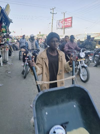 The image depicts a bustling street scene in Kwekwe, Zimbabwe, under clear daylight conditions. A young male, likely an adolescent, is centered in the immediate foreground, pushing a dark green wheelbarrow directly towards the viewer. He wears a dark knit cap, a dark undershirt, and a light brown overcoat; his expression is neutral, and he looks straight ahead. The wheelbarrow's basin contains light-colored granular material and a small, white, circular object with dark markings. A light-colored rope is secured horizontally between the wheelbarrow's handles.
The middle ground features a paved road populated by multiple adult males and motorcycles. To the left, a market stall displays various packaged goods and produce. An adult male in a light blue shirt and cap is operating a motorcycle with a sidecar attachment, moving from left to right. Another adult male in a dark maroon shirt and cap is visible on a motorcycle to the right, behind the young man. Numerous other individuals and motorcycles are present, suggesting active traffic or a gathering.
The background shows urban infrastructure, including utility poles with overhead power lines. A large red billboard on the right side displays text, including "Beeline" and "WE WE GET," alongside an image. Additional buildings and structures consistent with a town or market area are visible under a clear, light blue sky.