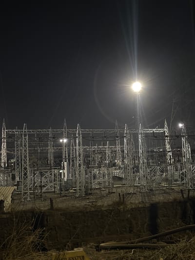 The image depicts an electrical substation in Ambernath, India, at night. A bright light source, possibly a street lamp or the moon, illuminates the scene. The substation is constructed of metal framework, with numerous electrical components and wires visible. A dark cliff or wall is situated adjacent to the substation. The sky is dark, and sparse vegetation is seen on the right side of the frame.
