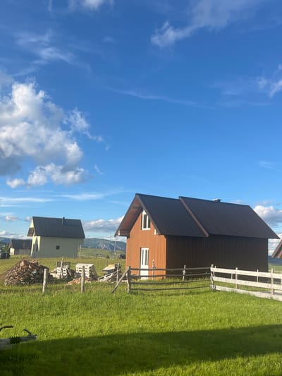 The image depicts a rural landscape under a bright blue sky with scattered white clouds. In the foreground, a neatly mown green lawn is visible, intersected by a rustic wooden fence. Behind the fence, several structures are present, including a prominent reddish-brown wooden house with a dark metal roof. To its left, a lighter colored building, possibly a barn or another house, also has a dark metal roof. In the middle ground, piles of wood and some wooden pallets are stored. The background features rolling hills and mountains, suggesting a location in a mountainous region. A dark-colored vehicle is partially visible on a road in the distance. The overall scene evokes a sense of tranquility and rural living.