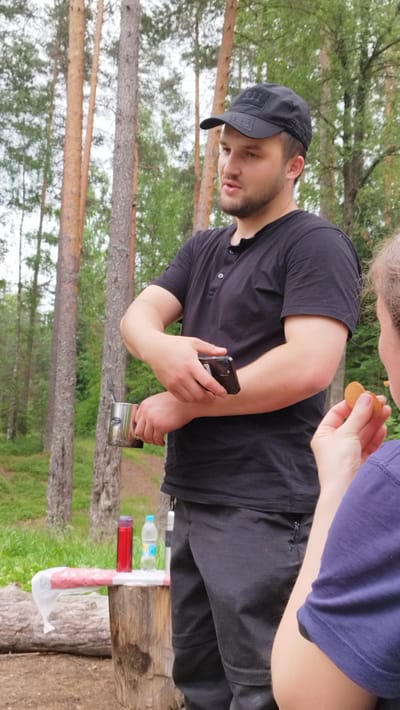 This image depicts a casual outdoor gathering in a forest setting, likely in Orekhovo, Russia. The primary subject is a young man wearing a black t-shirt, dark pants, and a black baseball cap. He is holding a smartphone in his right hand and a metal camping mug with a fork sticking out of it in his left. His gaze is directed off-camera, and his expression is neutral.
In the foreground, a portion of another person's arm and shoulder is visible, wearing a purple t-shirt. This person is holding a biscuit.
Behind the man, a wooden tree stump serves as a makeshift table. On it, there are two bottles: a red thermos and a clear plastic bottle of water. A white plastic bag with red stripes is also present. The background consists of tall pine trees, indicative of a forest environment. The lighting suggests it is daytime, and the lush green foliage implies a warm season. The overall mood seems relaxed and informal, possibly a picnic or a break during an outdoor activity. There is no visible text in the image, other than what might be on the water bottle label, which is too blurry to read.