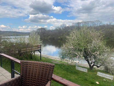A view of a river landscape in Smederevska Palanka Municipality, Serbia, from a patio or deck. In the foreground, a brown wicker chair is partially visible behind a brown wooden railing. The river dominates the middle ground, reflecting the sky and clouds. The opposite bank is lined with trees, some in bloom. A wooden deck extends toward the river. In the grass is a dilapidated white fence. The sky is filled with white clouds against a blue background.