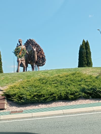 The image displays a monument situated on a landscaped island within a roundabout in El Espinar, Spain, under a clear, light blue sky. The central feature is a bronze-toned metal sculpture depicting a male figure, dressed in a hat and tunic, holding reins while standing beside a pack animal, identified as a donkey or mule. The animal carries a large, stylized load composed of angular metal pieces resembling stacked logs. A green wreath adorned with red and yellow ribbons is draped around the human figure's neck.
The sculpture rests on a gently sloped, grass-covered mound. Tall, slender evergreen trees are situated around the mound; one is partially visible on the far left, and a prominent cluster of three stands on the far right. A dark street light fixture is partially visible in the upper right of the frame.
In the immediate foreground, a grey asphalt road surface with a white painted line is present. This is bordered sequentially by a concrete curb, a section of paving with diagonal green and grey stripes, and a narrow strip of light-colored gravel. These elements precede a dense, low hedge of green shrubs forming the base of the grassy island. No active human presence or interaction is observed in the scene.