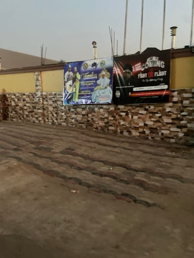 Two posters are affixed to a wall with patterned brickwork. The upper poster displays "HOME COMING FIGHT OR FLIGHT" in bold text, while the lower poster advertises an "Attestation Ceremony" for Chief Johnson C.N. Anagolu. The foreground shows a section of the patterned brickwork on the floor.
