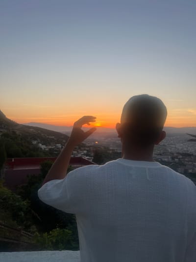 A person, seen from behind in silhouette, stands against a vibrant sunset overlooking the city of Tétouan, Morocco. The individual wears a light-colored, short-sleeved top with a subtle patterned knit. Their right arm is extended forward, with their open hand positioned to seemingly cup or frame the bright orange and yellow sun as it dips below the distant mountain horizon.

The sky exhibits a beautiful gradient, transitioning from the intense warm colors of the sun at the horizon to a soft blue-grey higher up. In the background, the silhouettes of mountains or hills are clearly visible, with the sprawling urban landscape of Tétouan nestled among them. Sparse buildings and structures can be discerned in the distant city. The foreground features dark green foliage, possibly from trees or bushes, suggesting an elevated viewpoint such as a balcony or terrace, with a hint of a white railing on the far right. The time of day is clearly sunset, creating a peaceful and contemplative atmosphere. There is no visible text in the image.