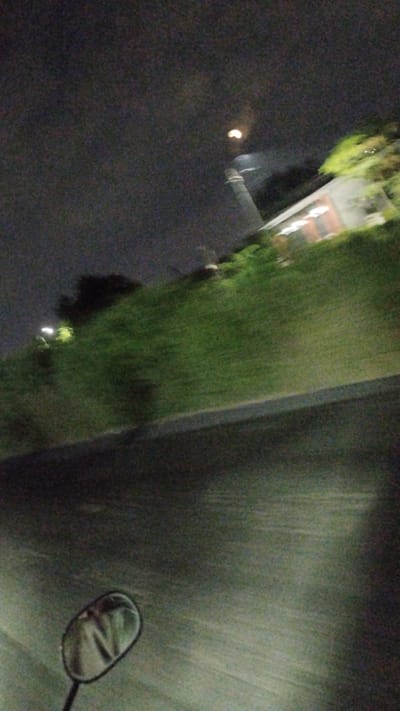 The image depicts a blurry night scene, likely captured from a moving vehicle such as a motorcycle, given the presence of a side-view mirror in the lower left corner. The mirror reflects a distorted image, possibly of the rider. The environment appears to be a road with a grassy embankment on the right side. A building with illuminated windows is visible on the embankment, and trees are present. A bright, circular object, possibly the moon or a street light, is visible in the dark, cloudy sky. The overall impression is one of low light conditions and motion blur, suggesting a nighttime journey. The location is identified as Sambungrejo, Indonesia.