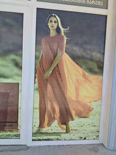 A full-body photograph of a woman in a long, flowing peach-colored dress stands in the middle of a sandy outdoor environment. The woman is facing forward with her head slightly tilted to her left and her gaze directed towards the viewer. Her hair is blowing to her left, suggesting wind. The dress has a fitted bodice with pleated details and a voluminous skirt that drapes around her, with a large portion of the skirt billowing to her right. Her bare feet are visible, one slightly forward. The background is a blur of muted greens and blues, possibly representing a distant landscape. To the left of the woman, a section of a product advertisement is visible, featuring the brand "AHAVA" and text that appears to be in a foreign language, with the words "APPLE OF SODOM" also present. The entire scene is framed by white borders, suggesting it is a display, possibly in a shop window. The photograph appears to be taken outdoors, with natural lighting. The presence of the "AHAVA" advertisement indicates the location is likely a commercial establishment. The text above the image, in Georgian script, suggests the image is located in Sartichala, Georgia.