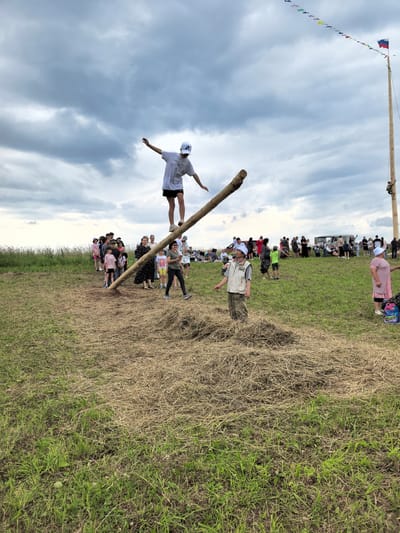 In Кояново, Russia, a young person wearing a white baseball cap and a white t-shirt with black shorts is balancing on a slanted log. They are in the center of the frame, with arms outstretched for stability. A crowd of people is gathered on a grassy field, observing the event. In the background, a tall wooden pole is erected, adorned with colorful triangular flags, and topped with a Russian flag. Several individuals are visible on the pole, with one person climbing. The environment consists of a mowed grassy field with patches of dry grass and hay. The sky is overcast with dramatic clouds.