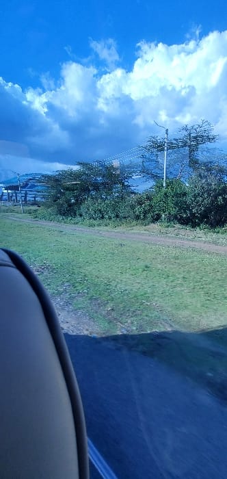 The image depicts an outdoor landscape within Hells Gate ward, Kenya, viewed from the interior of a vehicle. The top half of the frame shows a clear blue sky with scattered cumulus clouds, some appearing denser and shadowed. Below the sky, a verdant grassy area extends across the mid-ground, bisected by a narrow, unpaved dirt track. Further back, a line of various green trees and shrubs forms a natural barrier. Utility infrastructure includes a prominent utility pole with multiple horizontal lines extending from the right side towards the center. In the distant left, partially obscured by foliage, is a multi-story structure with a blue-hued roof or covering, accompanied by additional utility poles. Behind this, a calm body of water is visible, stretching towards a faint mountain or hill range on the horizon. The foreground's bottom-left quadrant shows the curved, light-colored upholstery of a vehicle's interior, likely a seat or trim. The bottom-right displays a dark, reflective surface, consistent with a vehicle's dashboard or a reflection on a window. No human or animal subjects are discernible within the frame.