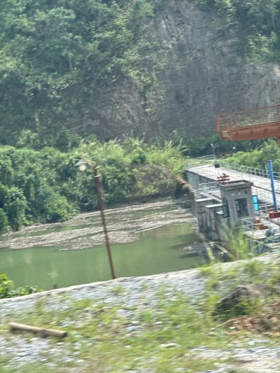This image captures a scene in Hoàng Su Phì Commune, Vietnam, featuring a river choked with debris. On the right side of the river is a gravel path with patchy grass growth. The river itself is a murky green, filled with what appears to be tree trunks and other wooden debris. On the left bank, a dense forest climbs up a steep mountainside. An industrial facility, possibly a hydroelectric plant, sits at the top edge of the riverbank, consisting of a concrete platform with structures and machinery. High-tension power lines are visible crossing over the river.