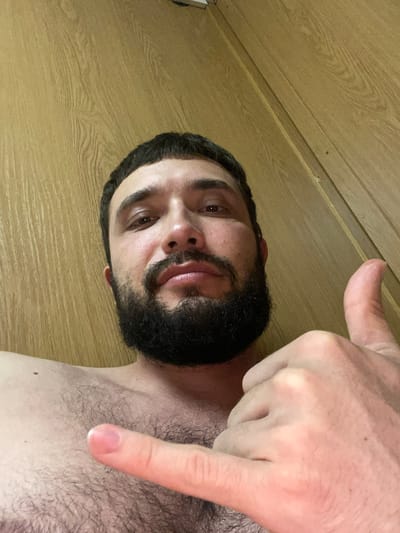 A man with dark hair and a beard is shown in a close-up, low-angle shot. He is shirtless, revealing chest hair, and is making a "shaka" hand gesture with his left hand and a thumbs-up with his right hand. The background consists of a wooden wall with visible grain and some seams. The lighting is warm and appears to be from an overhead source. No specific location in Bangladesh is identifiable from the image.