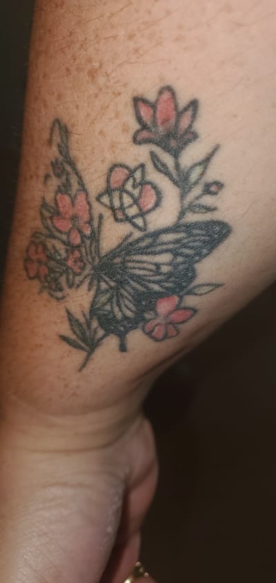 A close-up, low-angle shot shows a tattoo of a butterfly with pink flowers on a person's forearm. The tattoo is rendered in black ink for the butterfly and its floral companions, with accents of pink for the petals of the flowers. Some of the flowers appear to be in bloom, while others are buds. The butterfly's wings are detailed with intricate patterns. The skin of the forearm displays numerous small, brown freckles. The background is dark and out of focus. The image was taken in Sasbach, Germany.