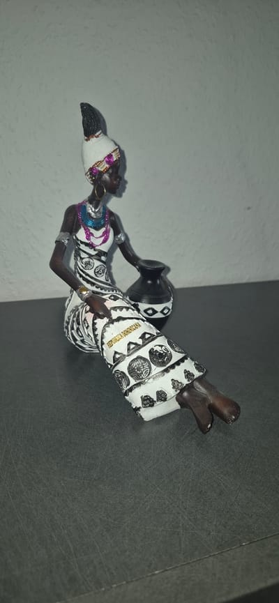 A figurine of an African woman in traditional attire is seated with a pot. The woman is dark-skinned with braided hair, wearing a white headwrap adorned with colorful beads and gold accents. Her dress is white with intricate black and silver patterns, and she has on beaded necklaces and earrings. She is holding a black pot with geometric designs. The figurine is placed on a dark, textured surface, with a light-colored, textured wall in the background. There is no indication in the image that this scene is located in Baden-Baden, Germany.