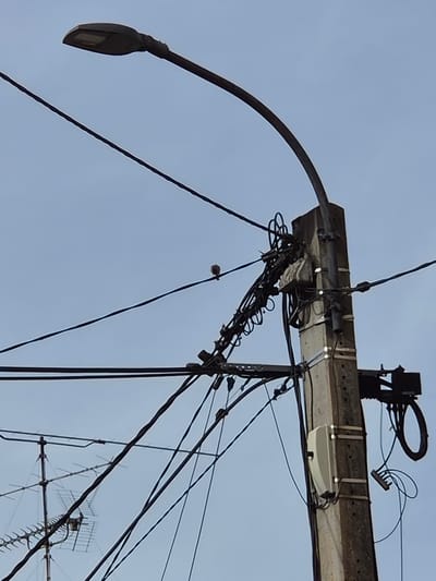 The image shows a utility pole against a pale blue sky. Attached to the pole is a curved streetlamp arm with a teardrop-shaped LED light fixture at its end. A rectangular, possibly wooden, platform is mounted atop the pole, hosting various electrical components, including junction boxes, wiring, and a white, rectangular device. Numerous wires and cables radiate from the pole and platform in multiple directions. A television antenna is visible to the lower right of the pole. The image originates from Audun-le-Tiche, France.