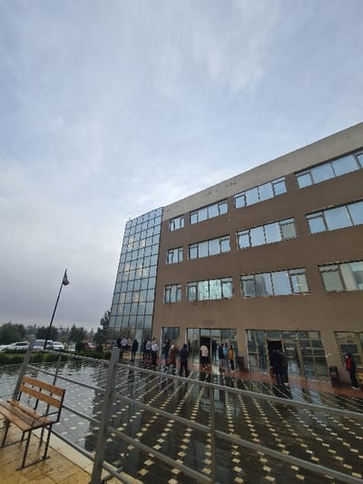 A tall, tan building with numerous windows dominates the view. The lower levels feature a wall of glass windows. People are gathered near the entrance under a patterned, covered walkway. A street lamp stands in the lower part of the frame, and a bench sits to the right. The sky above is overcast, suggesting a rainy or cloudy day.