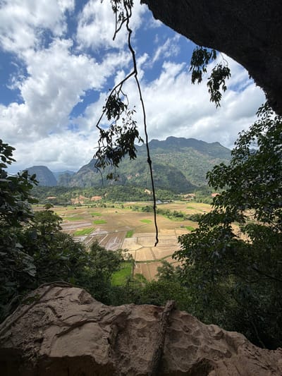 This image depicts a scenic view from a rocky overhang, looking out over a valley in Ban Nampoun, Laos. The foreground is dominated by a large, rough rock formation, with dark green foliage and branches framing the view. Beyond this, the landscape unfolds into a patchwork of agricultural fields, some appearing to be flooded for rice cultivation and others dry and harvested, in various shades of brown and green. In the distance, rolling, verdant mountains rise, some shrouded in clouds. The sky is a bright blue, dotted with fluffy white clouds. The scene is one of natural beauty and rural tranquility.