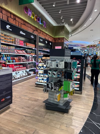 This image depicts an interior scene within what appears to be an airport duty-free shop, likely located in Cancún, Mexico. The store features a wide array of merchandise displayed on shelves and racks. Dominant products include confectionery items, such as chocolates, and electronic accessories like earbuds and power banks. A prominent "CHOCOLATE" sign with colorful lettering is visible at the top of the store, along with branding for "Folgado" and "Turin". In the mid-ground, several plush teddy bears with "MEXICO" emblazoned on their chests are displayed, along with other novelty items. In the background, other shoppers and employees are visible, with one employee in a green shirt with "Avola" printed on it walking away from the camera. The flooring transitions from wooden planks to polished black marble. The overall environment suggests a retail space designed to cater to travelers, likely at an international airport.