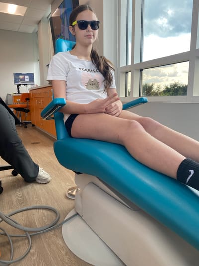 A young girl with long brown hair is reclining on a bright blue dental chair, wearing sunglasses and a white t-shirt with a cat graphic and the text "PURRFECT". She is wearing black socks with a white Nike swoosh. Her legs are bent, and her arms are resting on her legs. The setting appears to be a dental office, with cabinets and medical equipment visible in the background. A person's legs, wearing dark pants and white sneakers, are visible in the foreground, suggesting they are the person operating the camera or a dental professional. The scene is lit by natural light coming from a large window or skylight, showing a cloudy sky and some trees outside. The overall mood is casual and relaxed, possibly during a dental check-up or procedure.