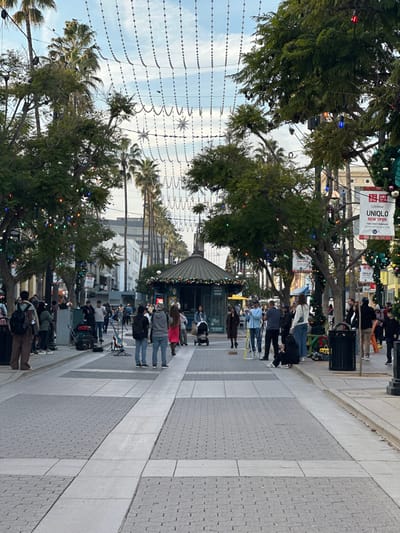 A street scene with numerous pedestrians populating a wide, patterned sidewalk. Trees line the street, adorned with Christmas lights. Overhead, decorative lights are strung across the street. Buildings are visible in the background. A kiosk-like structure is present in the middle of the walkway with pedestrians nearby.