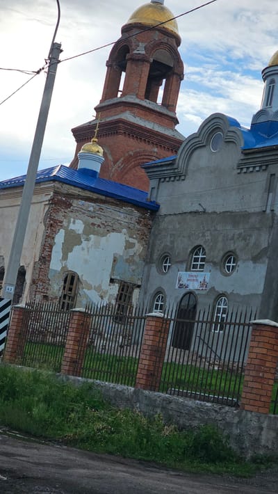 The image shows two adjoining religious buildings with blue roofs and golden domes. The building on the left appears older and in disrepair, with crumbling brickwork and missing plaster. It has arched windows and a smaller golden dome. The building on the right is more modern, made of grey concrete, and features arched windows with white cross-shaped panes. A banner with text and images of Easter eggs and a church is attached to the right building's facade. A brick and metal fence encloses the property, with lush green grass and vegetation growing behind it. A utility pole with wires stands to the left of the buildings, and a street runs in the foreground. The sky is cloudy with patches of blue. The location is identified as Pokrovskoye, Russia.