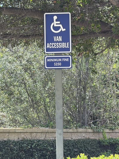 A portrait-oriented, vertical photograph displays two blue and white signs mounted on a gray metal pole, set against a backdrop of dense green foliage. The sign on the left features a white wheelchair accessibility symbol and the text "VAN ACCESSIBLE" in white capital letters. To its right, a second sign displays "MINIMUM FINE" in white capital letters, followed by "$250" in white numbers and a dollar sign. The signs are positioned in an outdoor setting, with sunlight filtering through the leaves of trees and bushes, suggesting a daytime scene. The lush vegetation, along with a hint of a paved walkway and manicured hedges to the right, indicates a public or semi-public area, possibly a park or a parking lot in Irvine, United States of America. There are no people visible in the image.