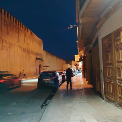 The image captures a street scene in Mechouar Fes Jdid, Morocco, at night. The street is lined with parked cars and an individual is standing on the sidewalk. On the left is a high, light-colored wall with battlements at the top. Across the street is a building with wooden doors and windows. A couple with a stroller is walking along the sidewalk on the left side of the street. A red car is moving in the foreground, slightly blurred. A streetlamp is positioned above, and the sky is a deep blue.